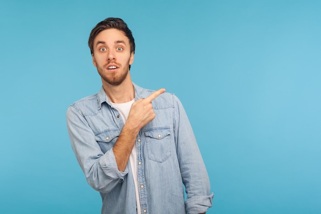 Wow, mira los anuncios aquí. Retrato de un hombre sorprendido con una camisa de denim apuntando a un lado, mostrando un espacio en blanco para la presentación de ideas, texto comercial. Foto de estudio interior aislado sobre fondo azul.