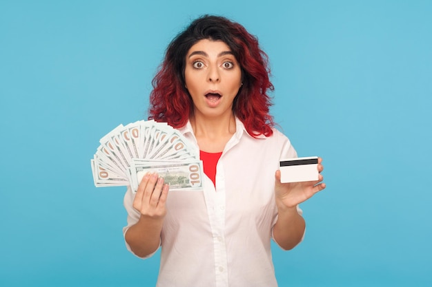 Wow, préstamo bancario para ir de compras. Retrato de una mujer hipster sorprendida con el pelo rojo elegante con tarjeta de crédito, billetes de dólar y mirando con la cara sorprendida. tiro del estudio aislado sobre fondo azul