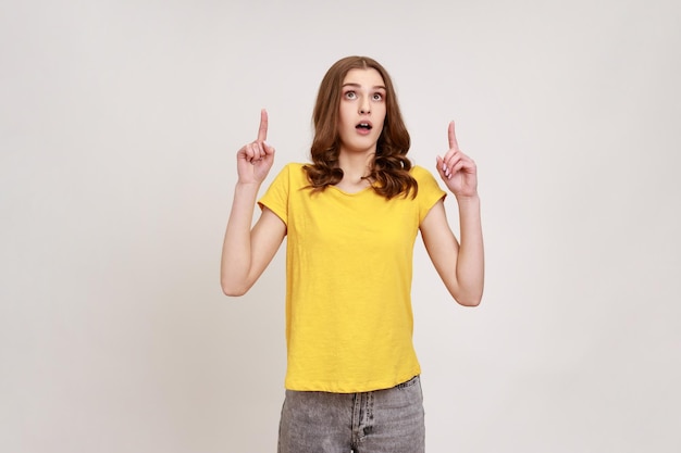 Wow, schau da Überrascht braunhaariges Mädchen in gelbem T-Shirt, das erstaunt aussieht und auf den Kopierbereich zeigt, der einen leeren Platz über dem Kopf zeigt Studioaufnahme im Innenbereich isoliert auf grauem Hintergrund