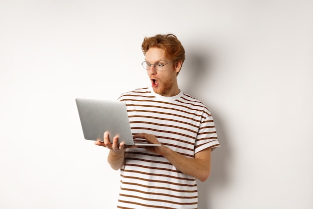 Wow, schau dir das an. Aufgeregter Rothaariger mit Brille, der auf den Laptop-Bildschirm starrt und Online-Promo-Angebot mit erstauntem Gesicht liest, weißer Hintergrund.