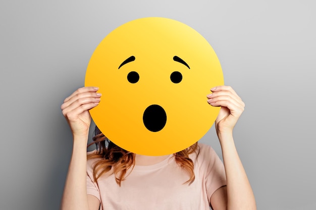 Wow schockiertes Emoji Mädchen hält einen gelben Smiley mit überraschtem Gesichtsemoticon