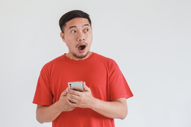 Wow und schockierter Gesichtsausdruck eines asiatischen Mannes, der ein rotes T-Shirt trägt und in den leeren Raum schaut, während er das Smartphone hält?