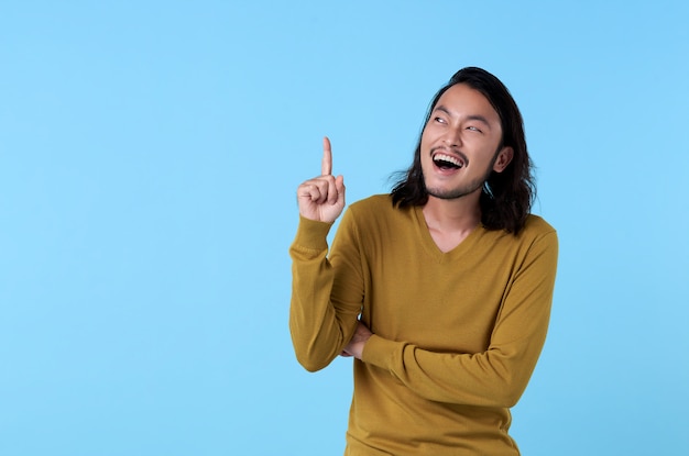 Wow und überraschter junger asiatischer Mann, der mit seinem Finger lächelt, der auf blauen Raum mit Kopienraum zeigt.