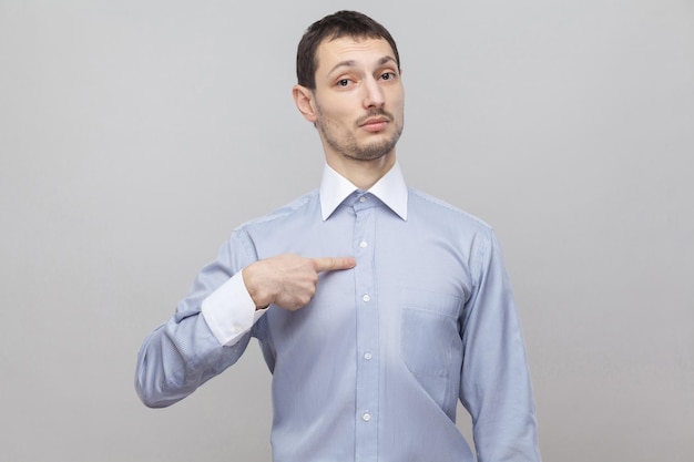 Foto wsou eu. retrato do empresário de cerdas bonito orgulhoso em pé clássico de camisa azul clara, apontando-se e olhando para a câmera. tiro de estúdio interior, isolado em copyspace de fundo cinza.