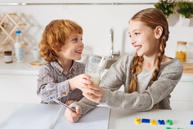 Würden Sie gerne einige. Charmanter freundlicher rothaariger Junge, der seinem Geschwister ein Glas Milch gibt, während beide den Morgen in der Küche genießen und am Tisch sitzen