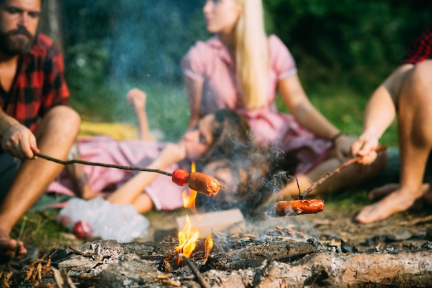 Würstchen auf Stöcken über den Flammen des Lagerfeuers kochen, im Sommer über Nacht campen. Gruppe von Freunden, die am Lagerfeuer sitzen