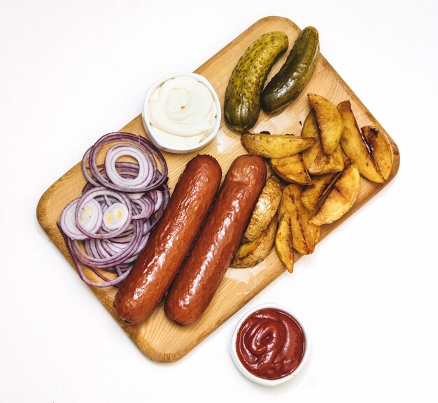 Würstchen mit Bratkartoffeln, Saucen, Zwiebeln und eingelegten Gurken auf Holzbrett auf weißem Hintergrund Draufsicht