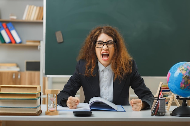 Wütend Blick in die Kamera Junge Lehrerin mit Brille sitzt am Schreibtisch mit Schulwerkzeugen im Klassenzimmer