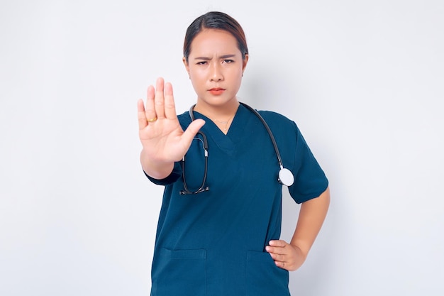 Wütend junge asiatische Frau Krankenschwester in blauer Uniform mit einem Stethoskop, das die Stirn runzelt und unzufrieden die Hand ausstreckt, um zu zeigen, dass das Stoppen nicht einverstanden ist, zu verbieten oder auf weißem Hintergrund zu verbieten Konzept der Gesundheitsmedizin