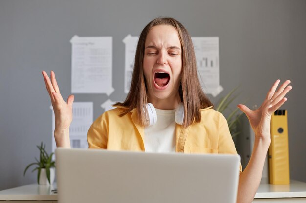 Wütende, aggressive, frustrierte junge Frau als Freiberuflerin arbeitete online im Heimbüro und blickte mit unzufriedenem, gereiztem Gesichtsausdruck und schreiend vor Hass auf den Laptop-Bildschirm