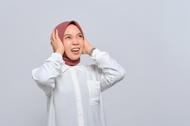 Wütende junge asiatische Muslimin schreit und bedeckt die Ohren mit beiden Händen, beunruhigt durch Lärm isoliert auf weißem Hintergrund