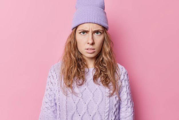 Foto wütende junge europäerin mit langen haaren hat einen gereizten gesichtsausdruck, grinst und sieht empört aus, trägt eine lila mütze und einen strickpullover und hört negative nachrichten oder schlechte worte, die isoliert über rosafarbenem hintergrund stehen