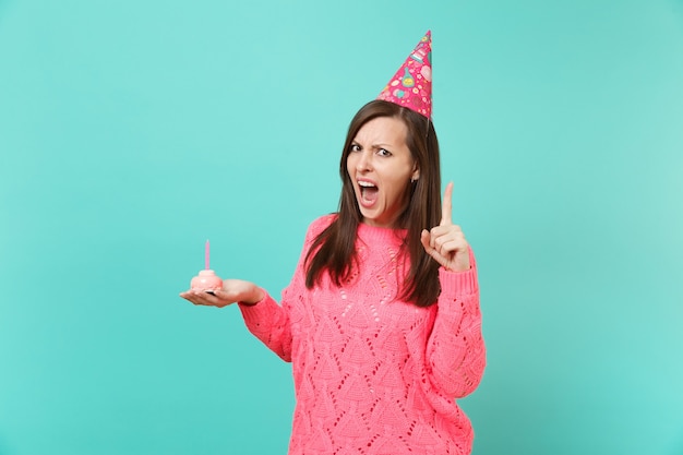 Wütende junge Frau in gestricktem rosa Pullover, Geburtstagshut schreien, Zeigefinger nach oben zeigend, Kuchen in der Hand haltend mit Kerze einzeln auf blauem Hintergrund. Menschen Lifestyle-Konzept. Kopieren Sie Platz.