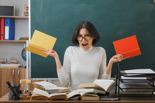 Wütende junge Lehrerin mit Büchern am Schreibtisch sitzend mit Schulwerkzeugen im Klassenzimmer