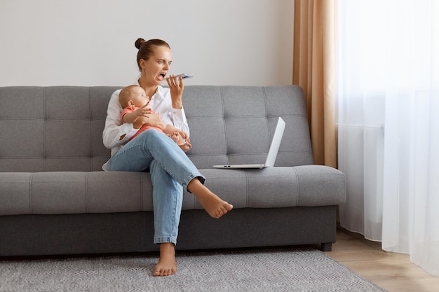 Wütende kaukasische Frau in weißem T-Shirt, die mit ihrer kleinen Tochter auf dem Sofa sitzt und an einem Laptop-Freiberufler arbeitet, der Probleme mit der Online-Arbeit hat, die schreiende Sprachnachrichten aufzeichnet