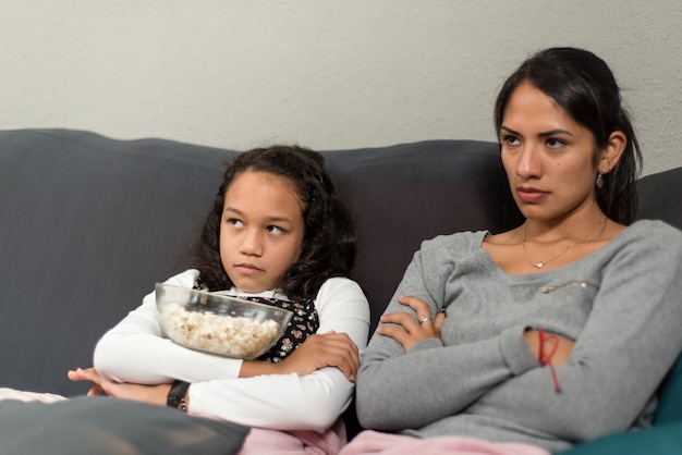 Wütende Mutter und Tochter mit verschränkten Armen, die zu Hause vom Sofa aus fernsehen.