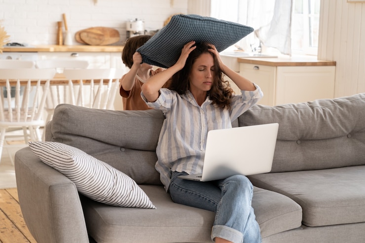 Foto wütende mutter versucht, von zu hause aus mit laptop auf der couch im wohnzimmer zu arbeiten, während der kleine sohn ihr kissen schlägt. schlecht benehmendes kleines kind nervt freiberufler remote worker mutter beim tippen von e-mails auf dem computer