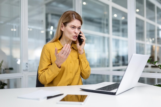 Wütende und verärgerte junge Geschäftsfrau sitzt im Büro am Schreibtisch und telefoniert