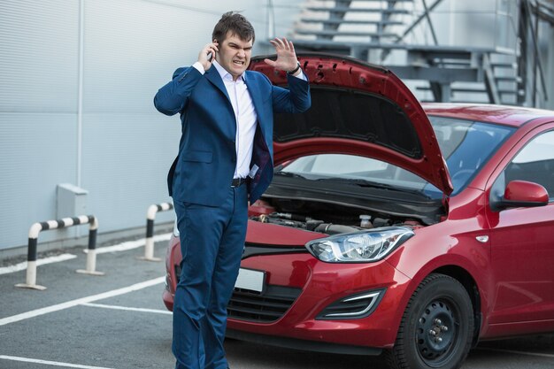 Wütender Geschäftsmann, der neben einem kaputten Auto steht und den Service anruft