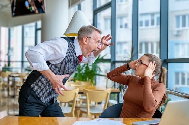 Wütender Geschäftsmann, der seine Unzufriedenheit mit der Arbeit an einem frustrierten Mädchen im modernen Büro ausdrückt.