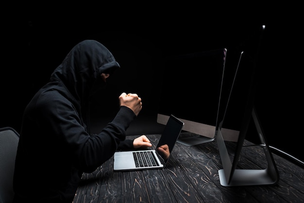 Wütender Hacker mit geballter Faust in der Nähe von Laptops und Computermonitoren mit isoliertem leerem Bildschirm