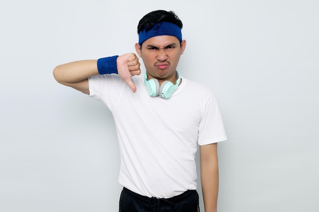 Wütender junger asiatischer Sportler mit blauem Stirnband und weißem Sportbekleidungs-T-Shirt mit Kopfhörern, die Ablehnung und Negativ mit Daumen nach unten zeigen, isoliert auf weißem Hintergrund Trainingssportkonzept