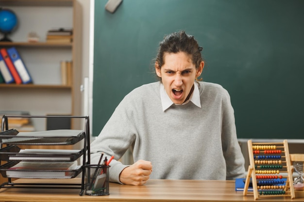 wütender junger männlicher lehrer, der die faust hält, sitzt am schreibtisch mit schulwerkzeugen im klassenzimmer