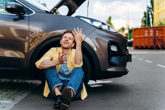 Wütender Mann, der mit offener Motorhaube am Steuer des kaputten Autos sitzt