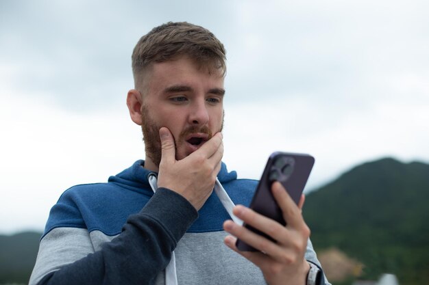 Wütender oder unglücklicher junger Mann, der auf den Bildschirm des Telefons im Telefon schaut, ist der Typ überrascht, aufgeregt und aufgeregt