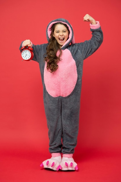 Wütendes Kind, das Schlafanzug trägt. Kigurumi-Pyjamas für Ihren Komfort. lustiges Kind im Hasenpyjama. Unterschiedliche Tageszeiten und Kinder planen das Konzept. Wecker am Morgen.