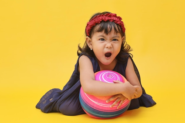 Wütendes kleines asiatisches Mädchen hält den Ball und sagt nein, während es über einem gelben Hintergrund sitzt