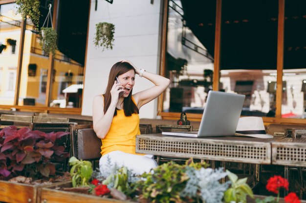 Wütendes Mädchen im Straßencafé im Freien, das am Tisch mit Laptop-PC sitzt, mit dem Handy spricht, schreit und Probleme stört, im Restaurant während der Freizeit