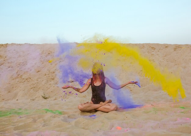 Wunderbare brünette Frau mit langen Haaren, die mit einer Wolke von Holi-Farbe posiert und auf dem Sand sitzt