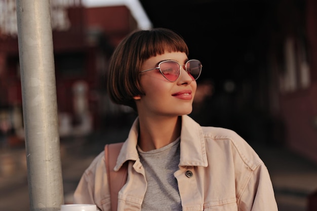 Wunderbare Dame mit kurzer Frisur, die draußen lächelt Schönes Teenager-Mädchen in heller Sonnenbrille, heller Jeansjacke und grauem T-Shirt, das auf die Straße blickt