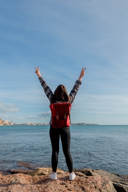 Wunderbare freudige junge Frau Reisende hebt ihre Arme glückliche Blicke auf das Meer im Freien an einem sonnigen Tag