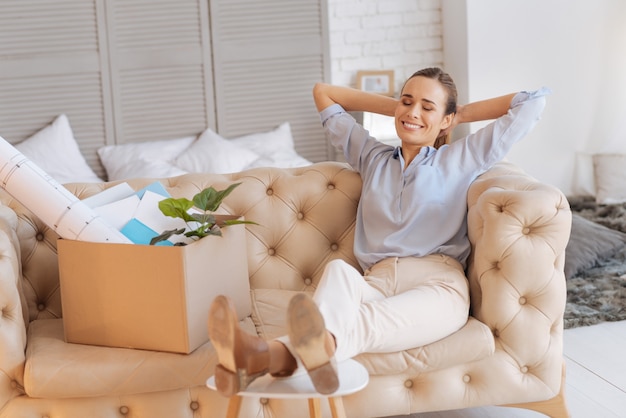 Wunderbare Stimmung. Fröhliche, positiv lächelnde Frau, die sich beim Entspannen auf dem Sofa nach dem ersten Tag bei ihrer neuen Arbeit gut fühlt