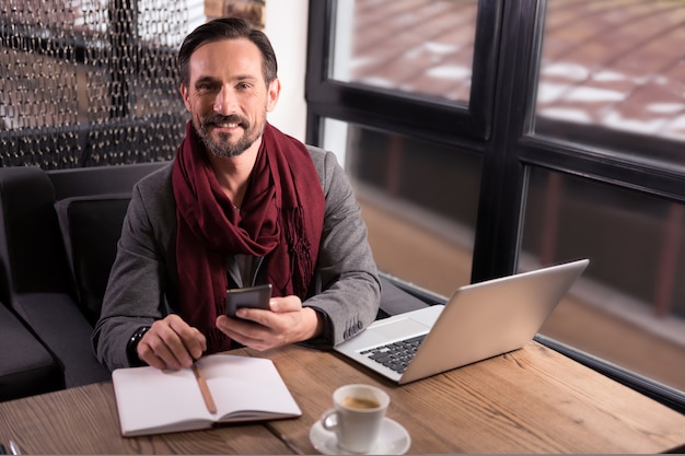 Wunderbare Stimmung. Positiver glücklicher netter Mann, der das Handy hält und Sie während der Arbeit im Café ansieht