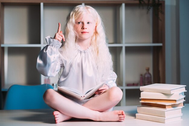 Wunderkind. Discovery-Einblick. Aha-Moment. Lernen zu Hause. Porträt eines inspirierten süßen blonden kleinen Albino-Mädchens, das mit offenem Buch auf dem Tisch sitzt und mit dem Finger nach oben zeigt.