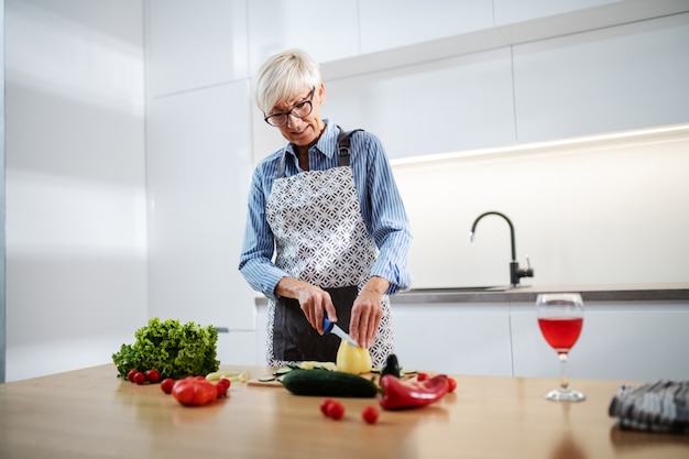 Wunderschöne ältere Frau in der Schürze, die Pfeffer einkauft und Salat macht, während er in der Küche steht