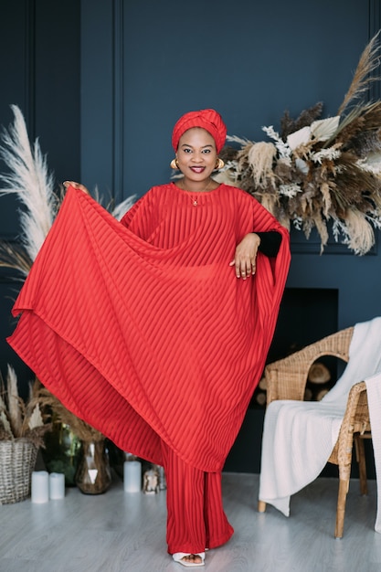 Wunderschöne afrikanische Frau in traditioneller roter Kleidung und Kopftuch, die im Studio mit rustikalen Boho-Dekorationen steht.