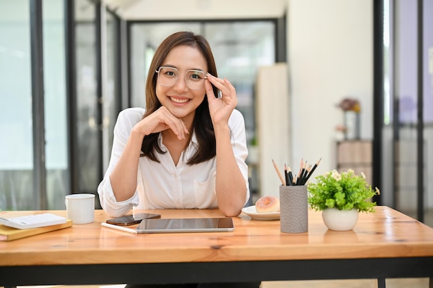 Wunderschöne asiatische Geschäftsfrau mit Brille lächelnd und an ihrem Schreibtisch sitzend