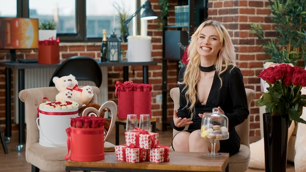 Wunderschöne Blondine im Videoanruf winkt der geliebten Person am Valentinstag zu und freut sich über Überraschungsgeschenke im Wohnzimmer. Schöne Frau, die lächelnd Hallo in die Kamera sagt und Geschenkboxen und rote Rosen genießt.