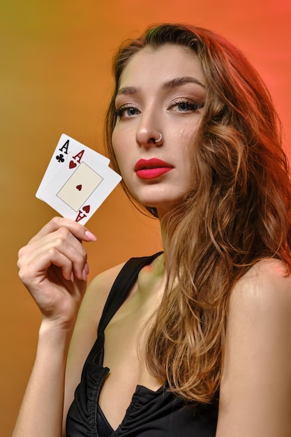 Wunderschöne brünette Frau mit hellem Make-up und Ohrring in der Nase, im schwarzen Kleid. Sie zeigt zwei Asse, während sie vor buntem Studiohintergrund posiert. Glücksspiel, Poker, Casino. Nahansicht