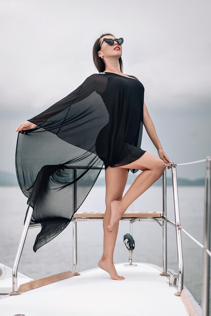 Wunderschöne Frau mit perfekter Figur in einem langen schwarzen Strandkleid und Sonnenbrille posiert auf dem Deck einer schneeweißen Yacht, Porträt