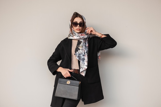 Wunderschöne hübsche junge Berufsfrau in stylischer Sonnenbrille im trendigen schwarzen Mantel mit Ledertasche mit einem Vintage-Schal auf dem Kopf, der im Freien in der Nähe einer Wand posiert. Business-Mädchen-Mode-Modell. Sexy Dame.