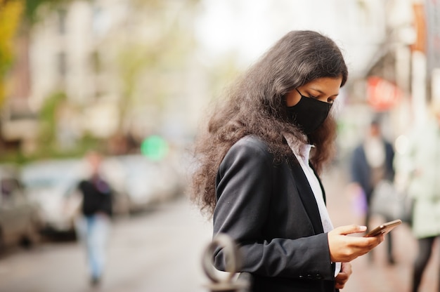 Wunderschöne Inderin tragen formelle und schwarze Gesichtsmaske, posiert auf der Straße während der Covid-Pandämie und sieht Nachrichten auf ihrem Smartphone.