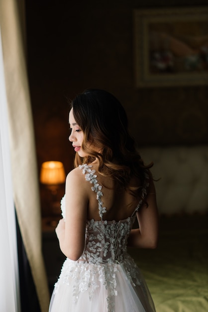 Wunderschöne junge asiatische Frau im eleganten Kleid, das Kamera und Fenster, Morgen der Braut, Hochzeit betrachtet