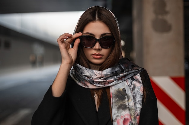 Wunderschöne junge brünette Frau in eleganter Kleidung richtet Sonnenbrillen gerade. Professionelles Mädchenmodell in einem modischen schwarzen Mantel mit einem Vintage-Seidenschal auf dem Kopf, der auf der Straße in der Nähe der Straße posiert.
