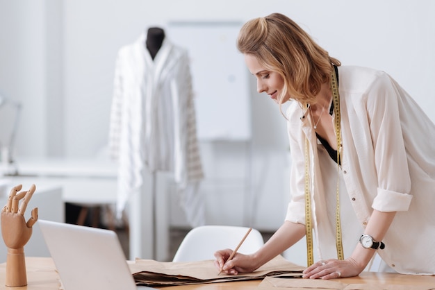 Wunderschöne junge Frau, die ein neues Kleidungsstück entwickelt und auf Mustern schreibt, die auf einem Tisch mit einem Laptop und einer Mannequinhand darauf liegen.