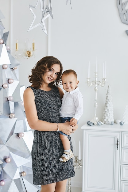 Wunderschöne junge Frau in einem modischen Kleid posiert mit ihrem süßen kleinen Sohn im weihnachtlich dekorierten Innenraum